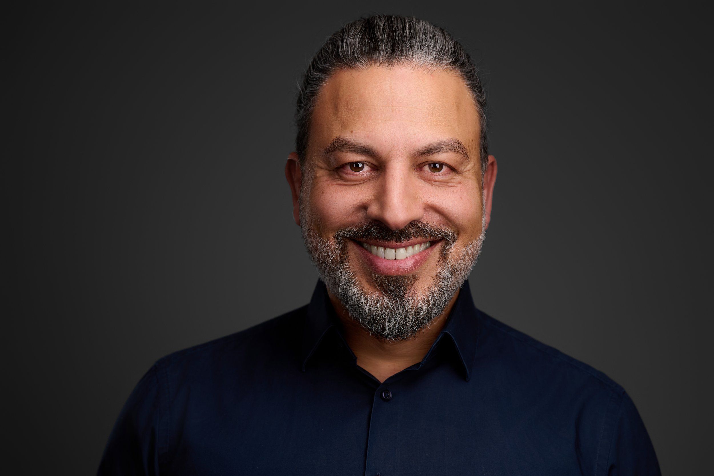 Headshot von Taylan: lächelnder Mann mit grauem Bart und dunklem Hemd vor dunklem Hintergrund