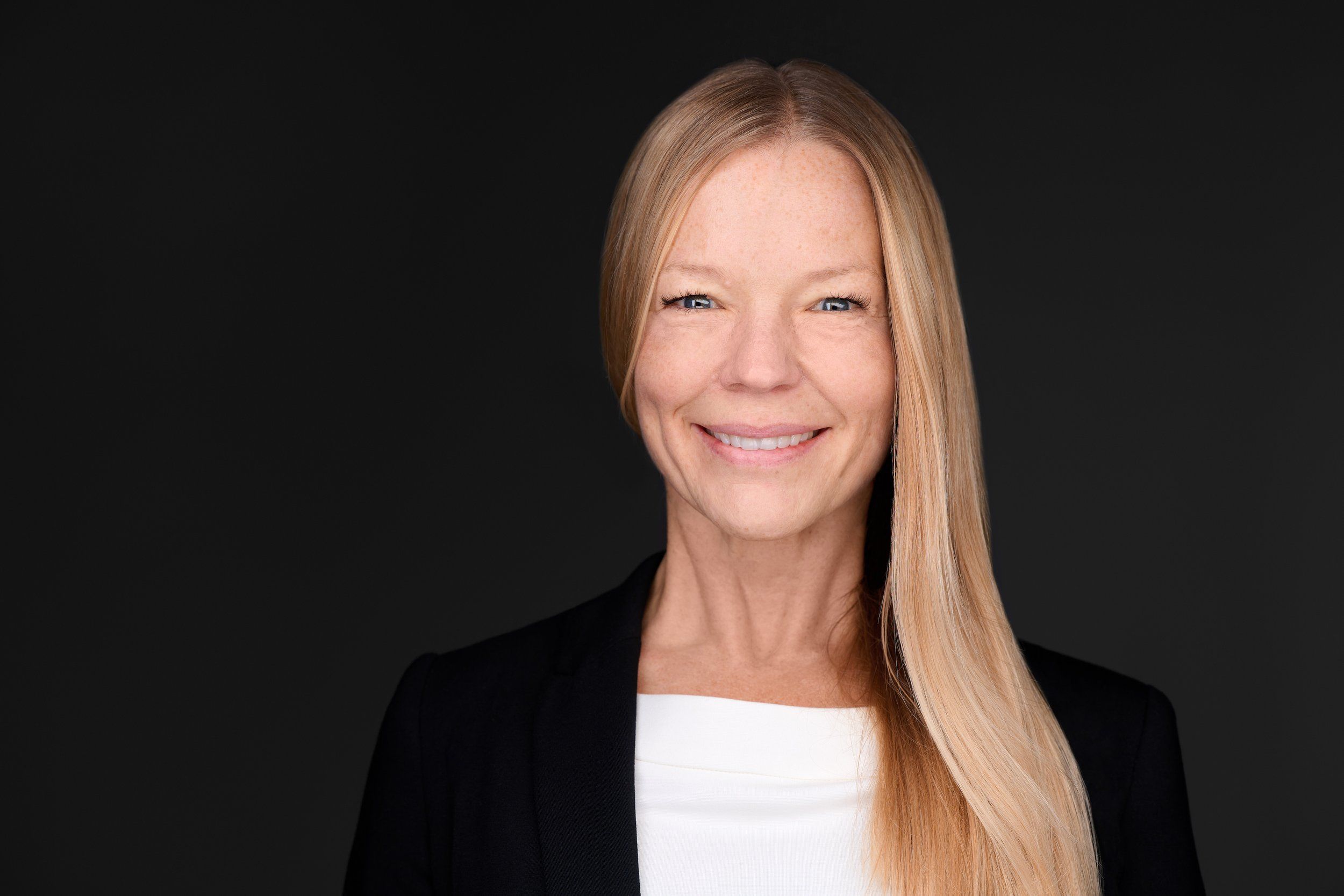 Headshot von Nora: lächelnde Frau mit langen blonden Haaren in Business-Kleidung vor schwarzem Hintergrund