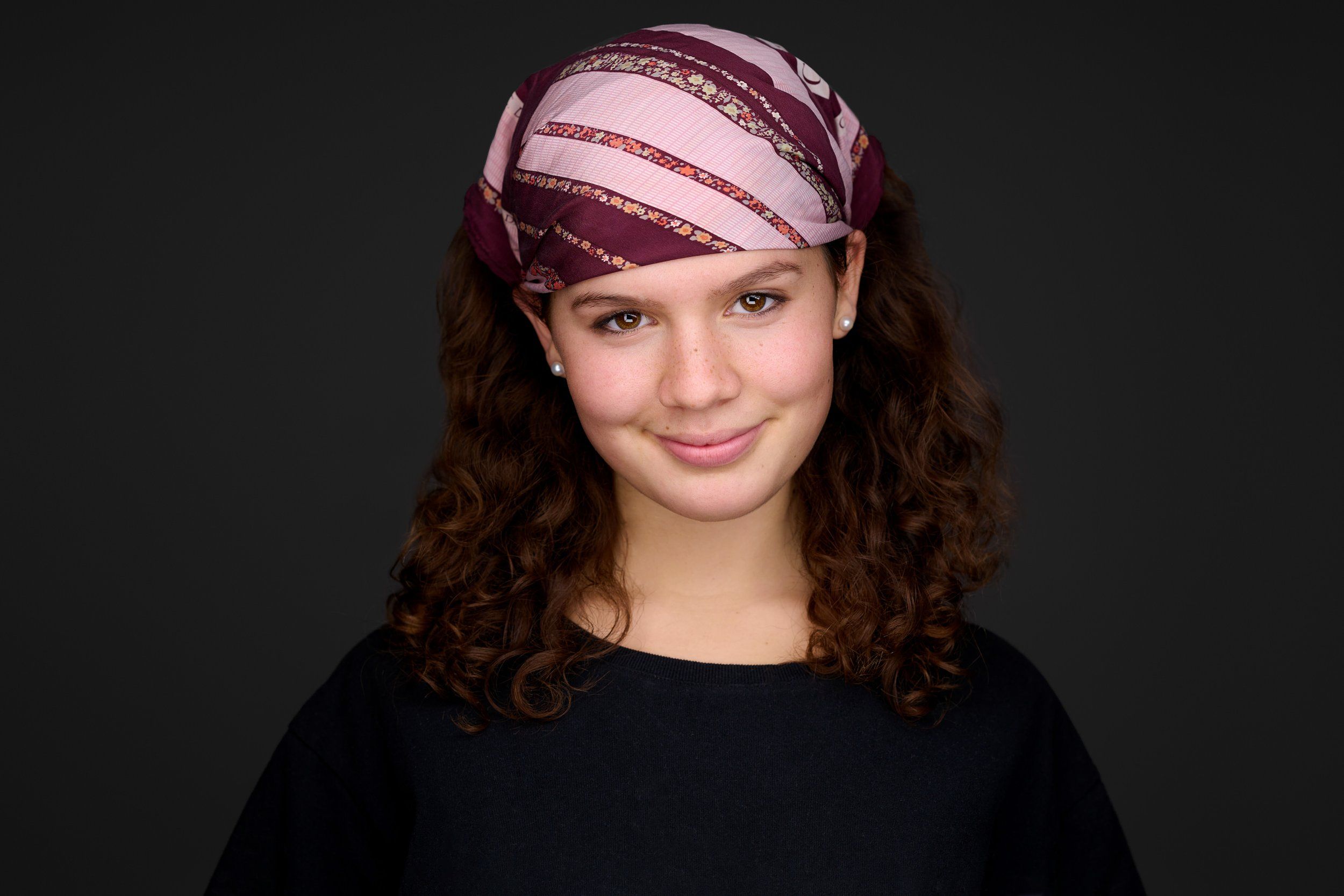 Headshot von Laura: junge Frau mit dunkelbraunen Locken, bunt gemustertem Kopftuch und schwarzem Oberteil vor schwarzem Hintergrund